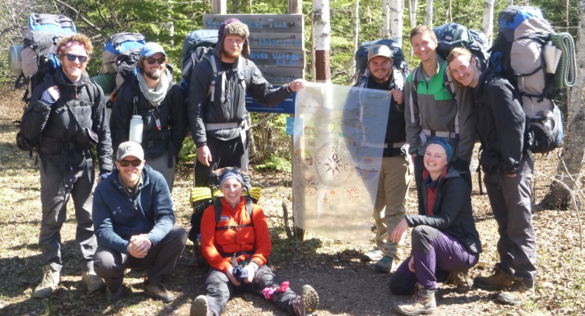 A group of students wearing backpacks pose for a photo with an Outward Bound sign. 